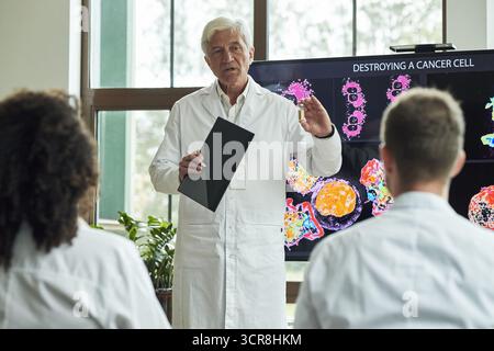 Senior Caucasian male doctor explaining cancer cell destruction process to diverse group of young colleagues in conference meeting, holding folder while standing in front of scientific display screen Stock Photo