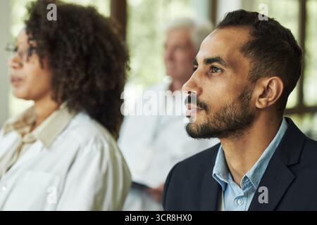Young Middle Eastern man listening attentively during seminar with American woman and senior Caucasian man in background, all focused on presentation or speaker Stock Photo