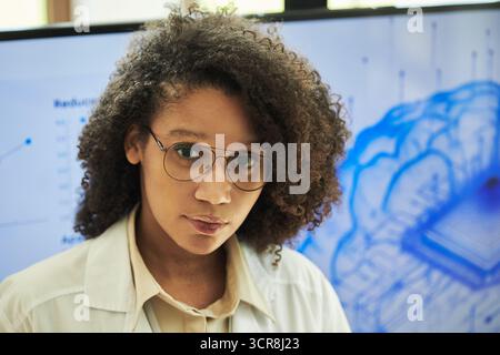 Portrait of young Black female scientist wearing white lab coat and eyeglasses standing in front of digital brain diagram display, showing confident expression, speaking at neurology conference Stock Photo