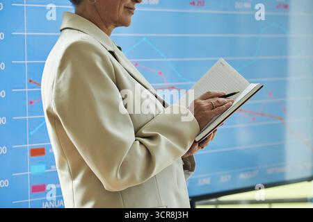 Middle aged Caucasian female conference presenter holding notebook and pen, standing in front of large digital financial chart, analyzing data, making notes, business professional in corporate setting Stock Photo