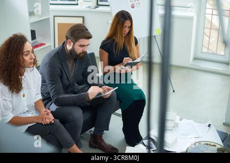 Young talented people sitting in modern office and waiting for discussion of new project. Asian woman making notes, bearded man in suit using tablet and curly woman looking at something pensively Stock Photo
