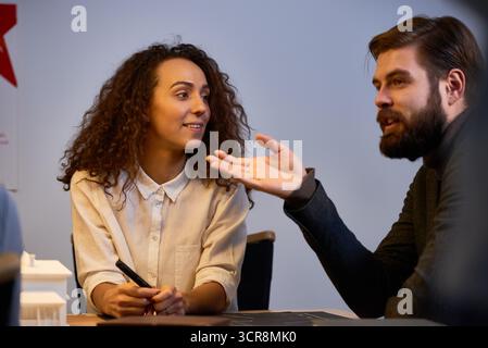 Young successful architects working on house project. Bearded man pointing at something and suggesting new idea while his curly female colleague looking at him with admiration and smiling Stock Photo