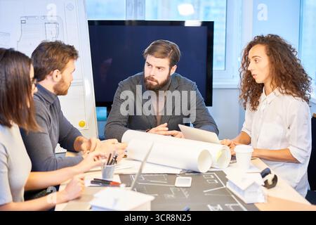 Team of designers discussing new project. Creative young people working on new apartment interior design, sitting at table covered with drafts and blueprints and sharing ideas Stock Photo
