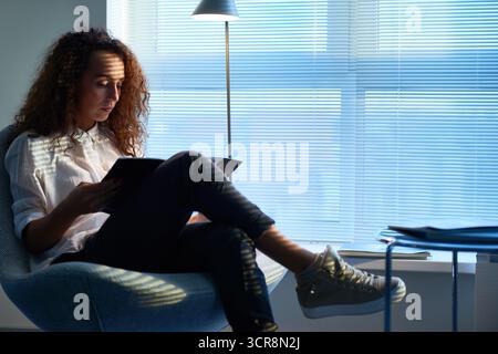 Young female stylist searching for new ideas reading fashion magazine. Curly woman sitting in soft chair with her legs crossed and leafing through pages while waiting for important clients Stock Photo