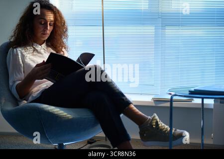 Pretty curly woman wearing white shirt and beige sneakers sitting in waiting room with Venetian blinds on windows, leafing through pages of fashion magazine Stock Photo