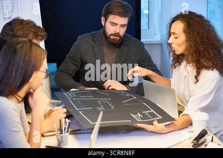 Stylish bearded chief architect discussing new house project with his colleagues. Attractive curly woman in white shirt presenting her draft made on black wove paper Stock Photo