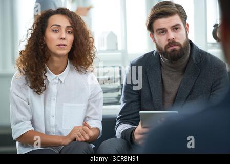 Concentrated curly brown-eyed woman sitting on office couch with clasped hands next to her bearded colleague in suit with tablet. Coworkers listening to important client attentively Stock Photo