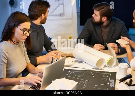 Professional architects having meeting at office, looking at new building draft attached to white board behind them. Concentrated young female specialist in round eyeglasses typing on laptop Stock Photo