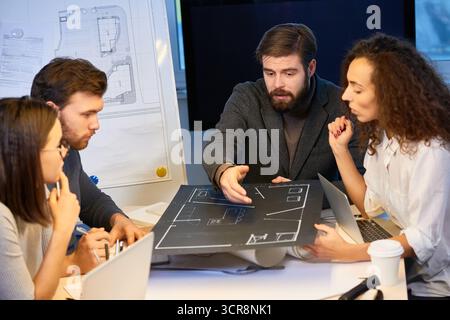 Experienced designers sitting at desk in office, holding new house layout made on black wove paper. Bearded chief pointing at draft and suggesting some corrections Stock Photo