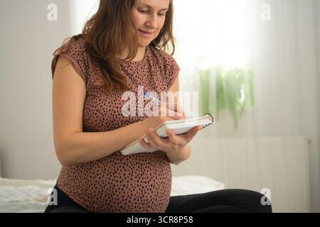 Pregnant woman holding pen and notebook, preparing a birth plan while sitting on bed. Stock Photo