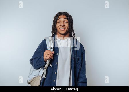 Young man with braided hair smiling and carrying backpack stands against a plain background Stock Photo