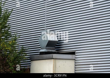 A gray ventilation unit is mounted on top of a white container beside a corrugated metal wall. Surrounding vegetation adds a touch of greenery to the industrial setting. Stock Photo