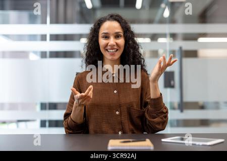 Young professional businesswoman smiling and gesturing while participating in an online video call, communicating and collaborating virtually from a modern office environment Stock Photo