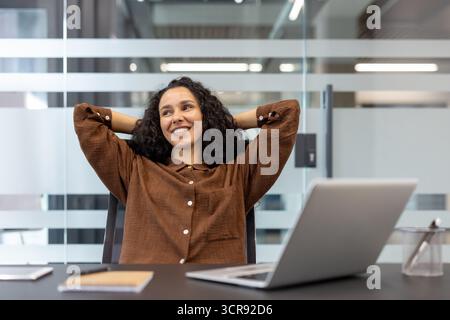 Young businesswoman with curly hair leaning back in her office chair, smiling and looking away from her laptop, enjoying a relaxed moment during a successful workday Stock Photo