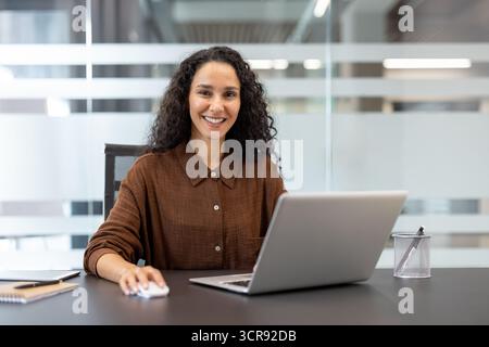 Young businesswoman with curly hair smiling at camera, using a laptop and mouse while sitting at a dark desk in a contemporary corporate office environment Stock Photo