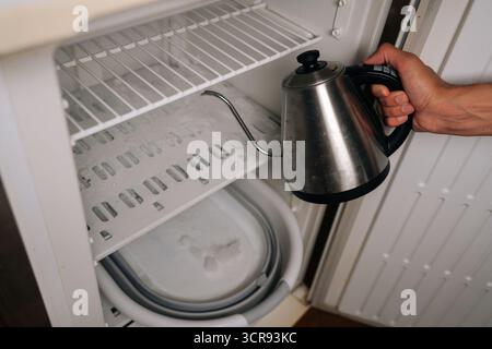 Close-up of unrecognizable person pouring hot water from kettle into refrigerator to melt ice and defrost, collecting melted ice in basin. Stock Photo