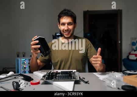 Portrait of positive computer technician installing CPU cooler on motherboard, working in dark workshop with scattered computer components, smiling Stock Photo