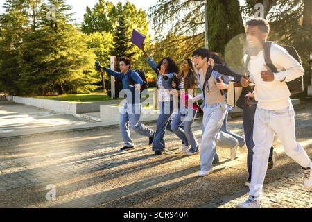 Group of excited multiracial students. Wearing casual clothes and carrying backpacks and books. Running across a university campus with trees in the b Stock Photo