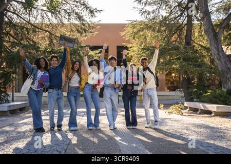 Group of diverse young university students happily standing in front of a campus building, smiling and waving, celebrating education, friendship, and Stock Photo