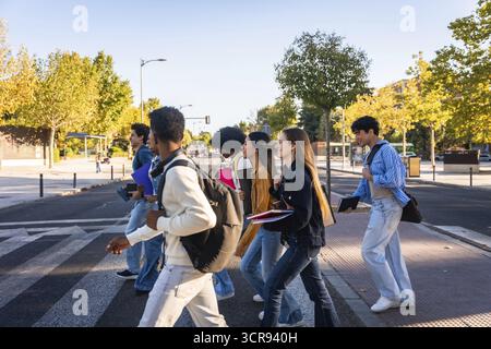 Group of diverse university students with backpacks and books walking across a pedestrian crosswalk, smiling and talking, representing education, frie Stock Photo