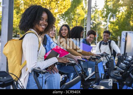 Group of happy multiracial university students smiling and preparing to use shared bicycles from a city station, promoting sustainable urban mobility Stock Photo