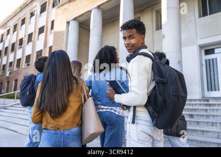 Group of diverse young university students with backpacks and books walking up the steps towards a campus building, representing education, friendship Stock Photo