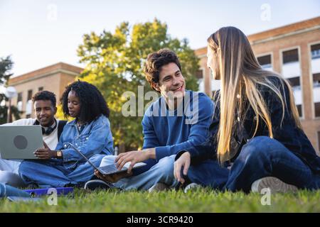 Group of diverse university students collaborating, studying, and engaging in conversations on the grass outdoors, using laptops and enjoying a break Stock Photo