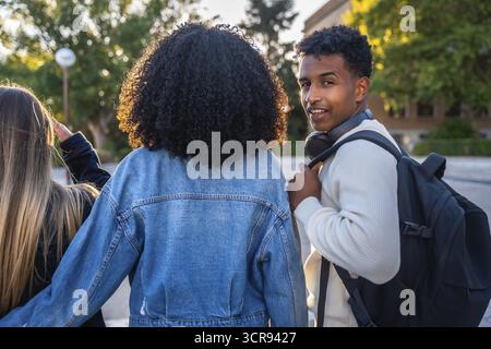 Three diverse young university students walking together on campus, the smiling man looking back at the camera while wearing headphones and a backpack Stock Photo