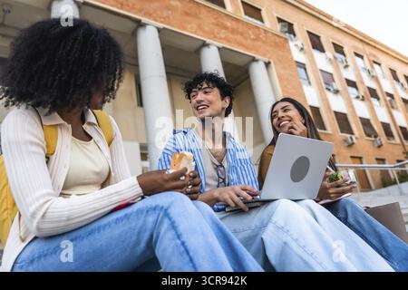 Group of happy young multiracial students sitting on steps outside a university building, enjoying a casual lunch and socializing between classes on c Stock Photo