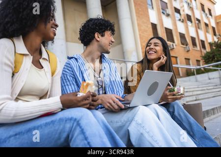 Group of diverse university students sharing a happy moment, laughing and eating lunch together on campus stairs, symbolizing friendship and student l Stock Photo