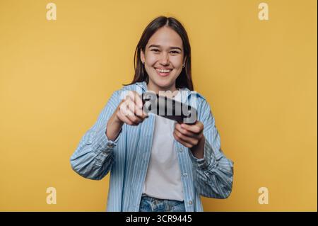 joyful young woman smiles while holding a game controller, embodying the fun and excitement of gaming on vibrant yellow background Stock Photo