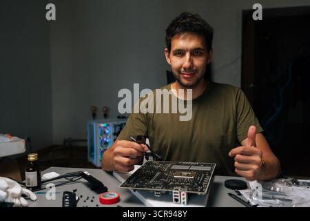 Smiling computer technician showing thumbs up gesture while fixing motherboard sitting at desk in workshop, surrounded by electronic components Stock Photo