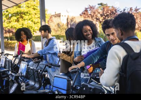 Group of happy diverse university students activating a shared bicycle from a station, preparing to ride on campus, representing sustainable urban tra Stock Photo