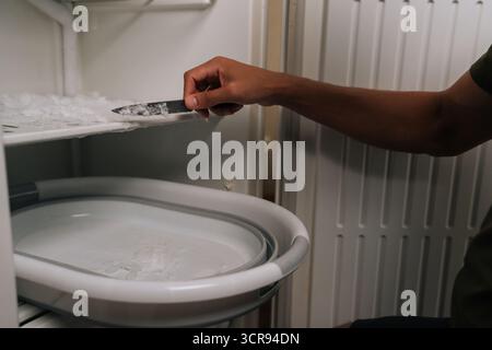 Close-up hands of unrecognizable man defrosting refrigerator, using knife to scrape off ice buildup and collecting ice chips in basin placed inside Stock Photo