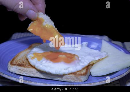 Close-up of a hand dipping bread into a runny egg yolk, served on toast with cheese, creating a delicious and simple breakfast Stock Photo