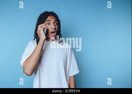 young man with dreadlocks talks on a smartphone, displaying astonishment and surprise on blue background Stock Photo