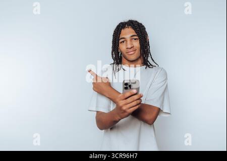 young man stands confidently pointing while holding a smartphone, reflecting a moment of direction and intention on white background Stock Photo