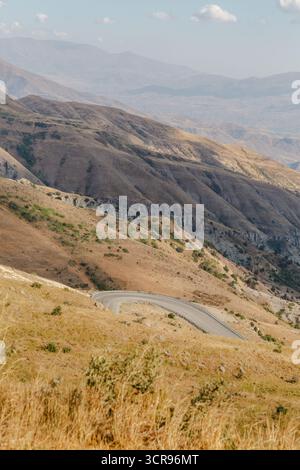 View of a winding road snakes through a sun-drenched, arid landscape, where golden grasses meet distant, hazy mountains, Vardenyats Pass, Armenia. Stock Photo