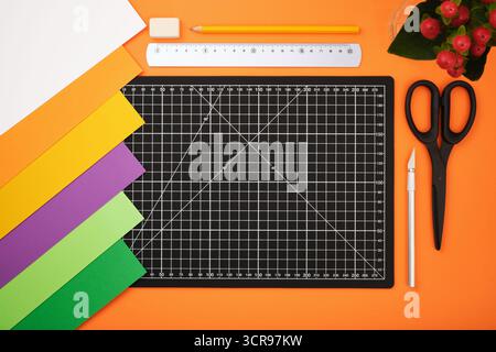 Colorful paper sheets, cutting mat, scissors, ruler, pencil, knife, and eraser on orange background. Desktop for paper crafts. Stock Photo