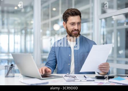 Young businessman smiling, holding paperwork while typing on laptop computer, managing finances and business operations in a modern office environment Stock Photo