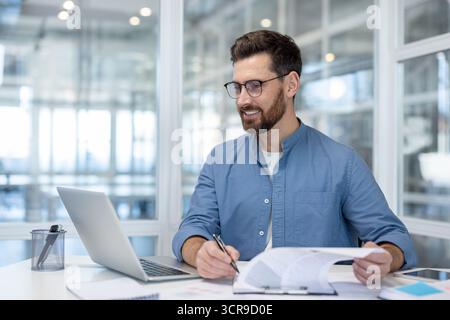Young businessman working confidently at his desk, reviewing documents and using a laptop with a positive expression, symbolizing productivity and career success in a modern workplace Stock Photo
