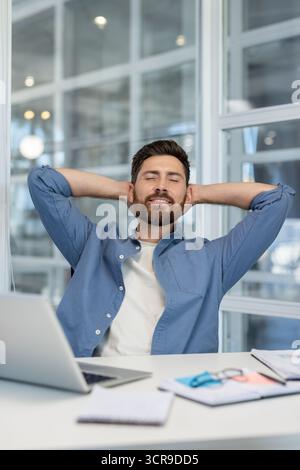 Smiling man finding serenity at his office desk, leaning back with hands behind his head, enjoying a moment of relaxation and satisfaction after achieving a task with his laptop nearby Stock Photo