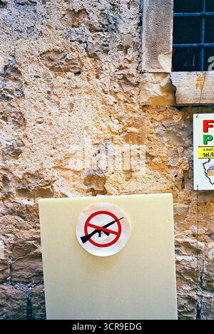 No weapons sign mounted on a board in front of a rough stone wall, photographed as a straightforward documentary-style analog snapshot. Stock Photo