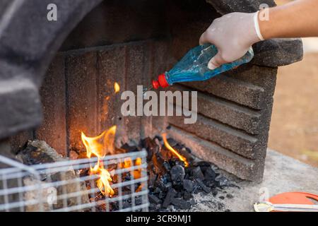 A gloved hand pours blue liquid onto burning charcoal in a stone barbecue grill. Flames rise from the coals, indicating preparation for cooking. Stock Photo