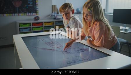 Schoolchildren Collaborate at Touchscreen, Examining Human Anatomy. Kids Talk, Actively Engage in Science Study. Concept of Modern Classroom Technology, Cooperative Biology Learning. Dolly Shot. Stock Photo