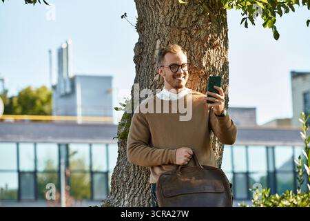 A young man in a sweater smiles at his phone by a tree on campus. Stock Photo