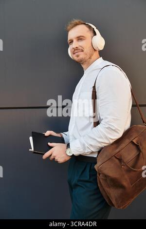 University teacher stands confidently against a wall, enjoying music with headphones and notes. Stock Photo