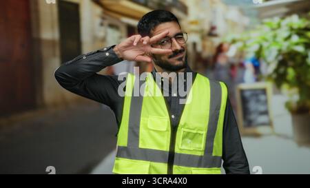 Young hispanic man in safety vest makes peace sign on urban street outside with smile. Stock Photo