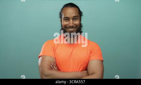 Man with crossed arms showing forearms, wearing bright orange shirt in teal studio; confidence resilience. Stock Photo