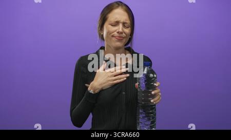 Young hispanic woman coughing into fist while holding large water bottle against a purple studio backdrop; dehydration discomfort. Stock Photo